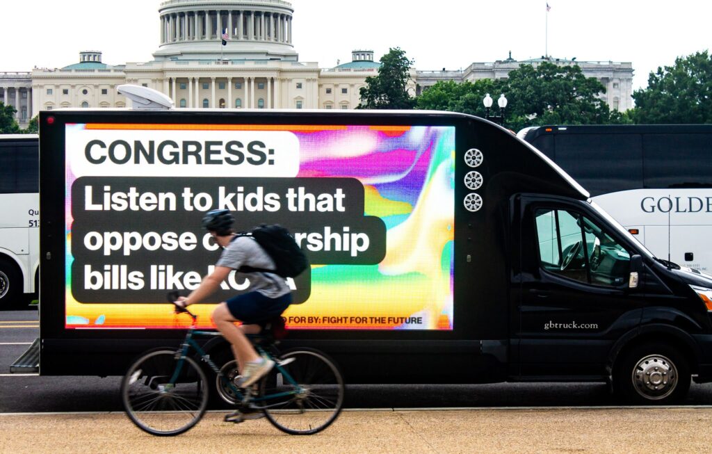 Exterior. Washington DC. A bicyclist rides by a truck displaying a digital billboard that reads "CONGRESS: Listen to kids that oppose censorship bills like KOSA"