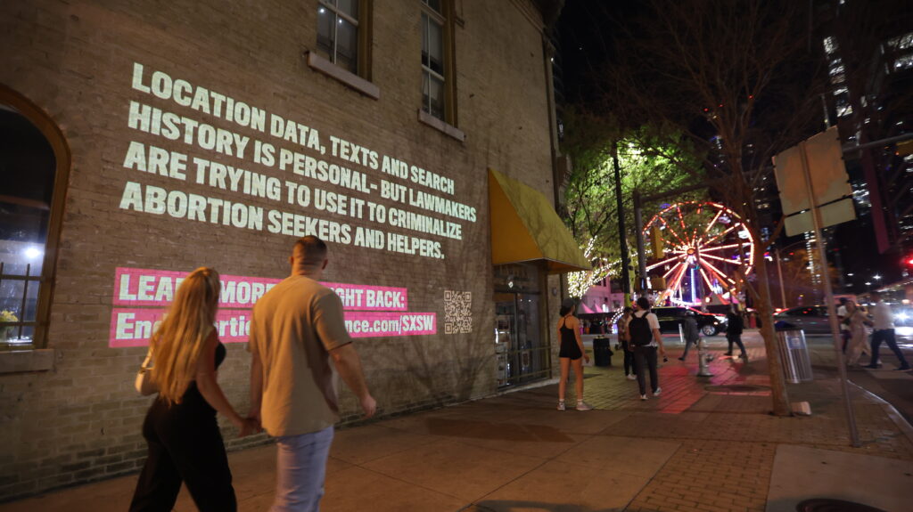 Several people walk by a projection that reads "Location data, texts and search history is personal - but lawmakers are trying to use it to criminalize abortion seekers and helpers." at South by Southwest.