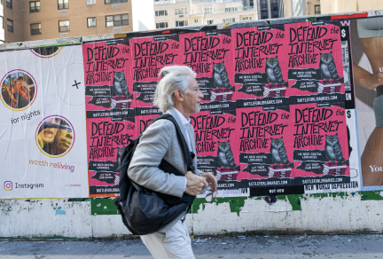 A person walks by a wall of wheat pasted posters that say "Defend the Internet Archive" in New York City.